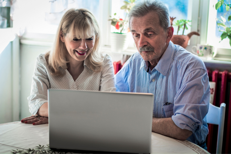 A photograph of an older man and a woman sat at a table looking at a laptop