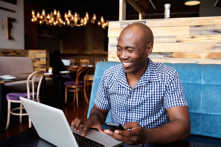 A photograph of a smiling man sat down using a computer in a café.