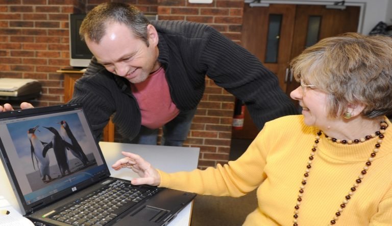 A photograph of an older woman sat at a desk using a laptop. Standing next to her, also looking at the laptop is a male digital volunteer. The screen of the laptop is displaying three penguins.