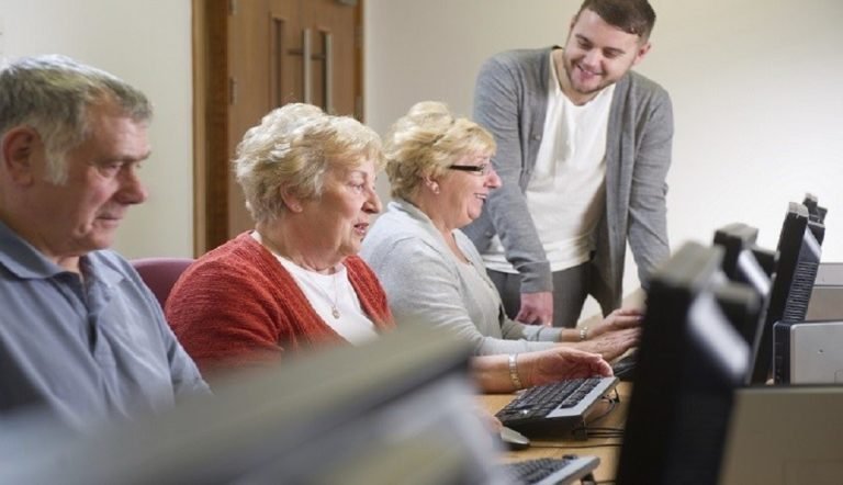 Two older women and a older man sit in a row behind a desk all looking at a computer screen each. At the end of the row is a younger man, who is a digital volunteer, that is stood up and smiling.