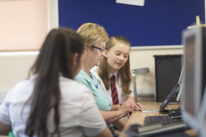 A photograph of an older woman sat at a desk using a computer. Either side of her are two female Digital Heroes, school pupils, who are helping her to use the computer.