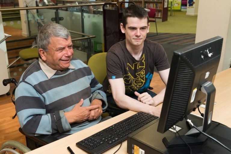 A photograph of an older man sitting next to a younger man, who is a digital volunteer. They're both sat at a desk in front of a computer while looking at the desktop monitor.