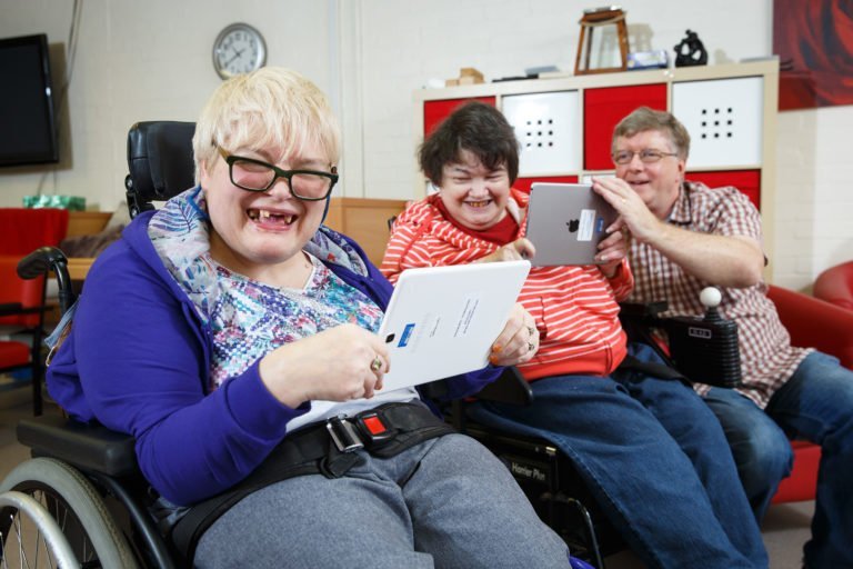 A photograph of two women in wheelchairs smiling for a picture while using tablets. There is a man in the background of the image who is sat on a chair and holding a tablet for one of the women.