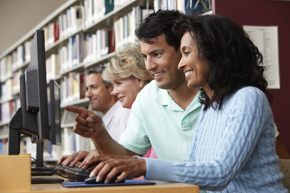 A photograph of two women and two men sat in a library at a desk using desktop computers.