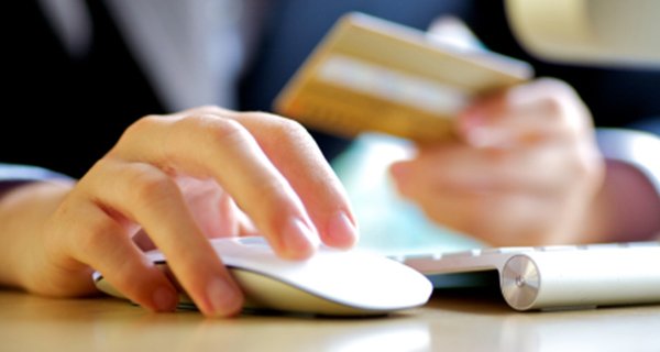 A close up of the hands of a person using a credit card to buy something online. Their right hand is on a computer mouse and their right hand is holding a bank card.