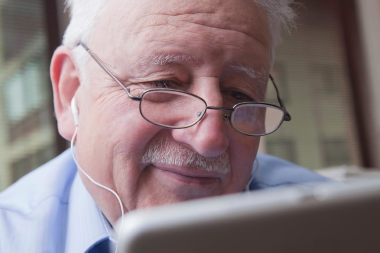 Close up photograph of a senior man wearing glasses with earphones in. He's smiling while looking down at a tablet device.