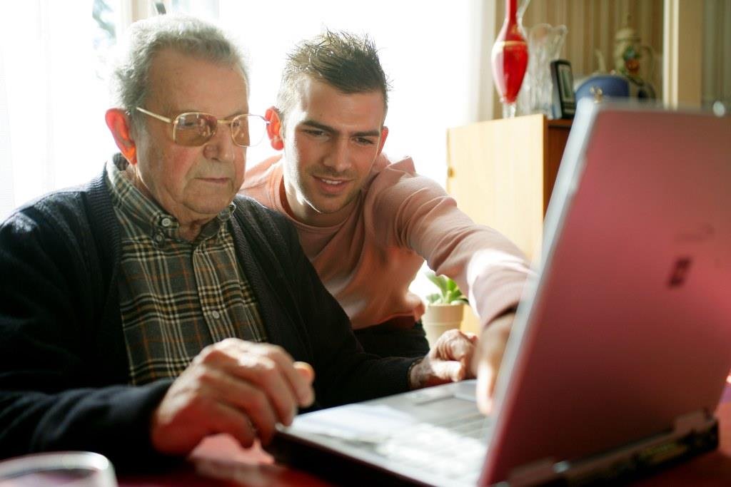 A photograph of a senior man sat down and looking at a computer screen. Squatting next to him is a younger man, a Digital Volunteer, who is pointing at the laptop screen.