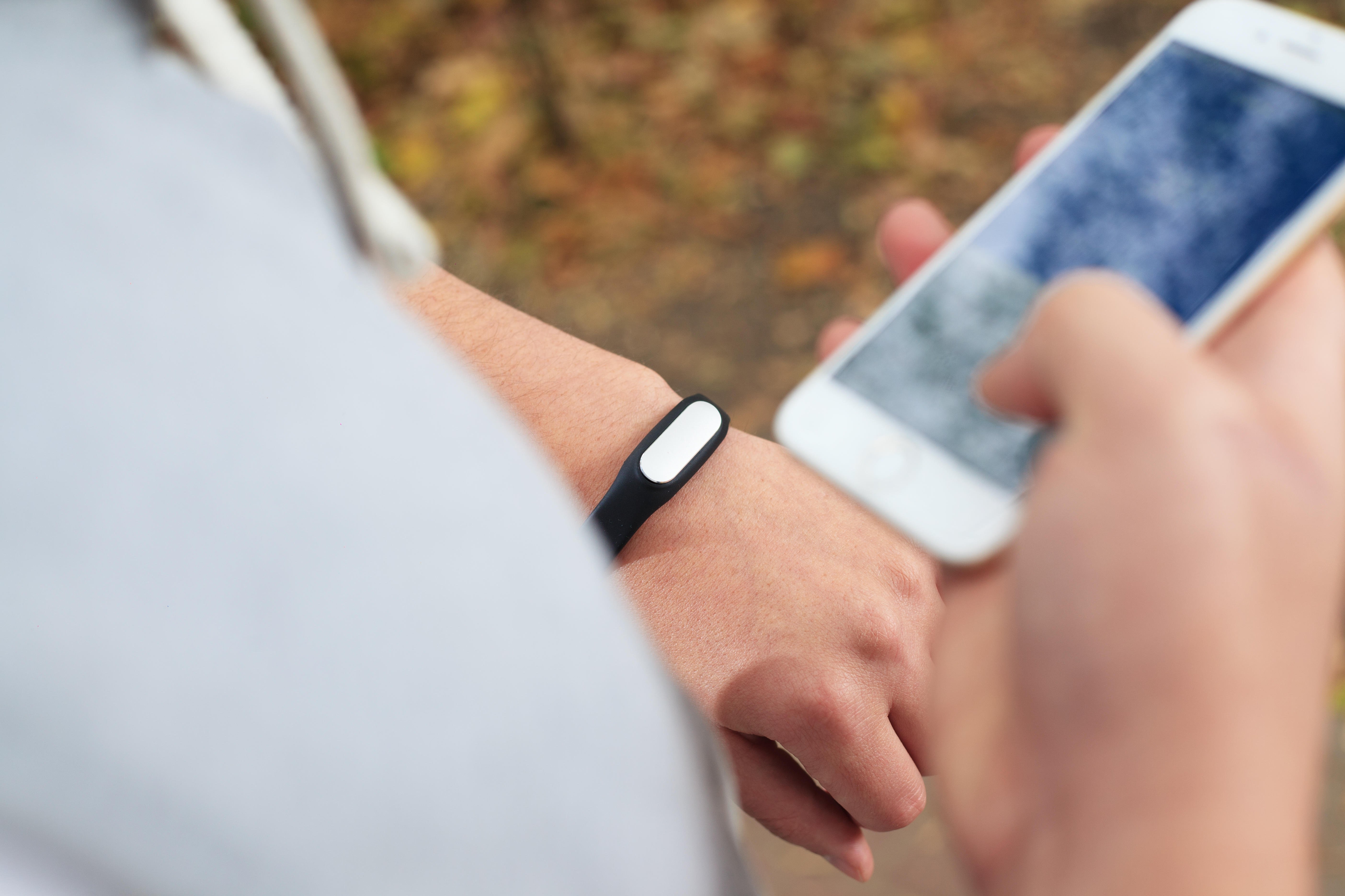 An over the shoulder photograph of a woman using a fitness bracelet. The bracelet is on her left wrist, while she holds an iPhone in her right hand.