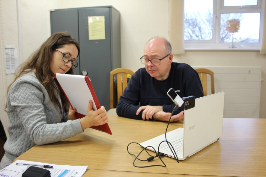 A photograph of a female staff member at Innovate Trust sat down at a desk, showing client how to use a tablet device, who is also sat at the desk. On the desk there is also a laptop.
