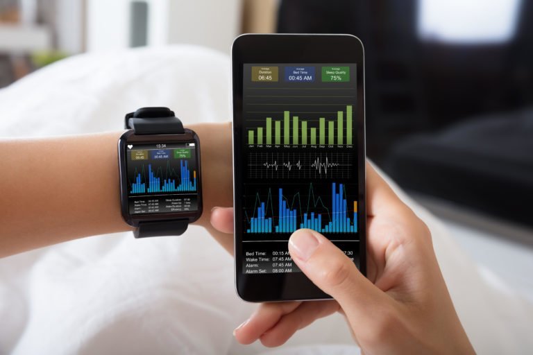 Close-up of a female right hand holding a mobile device with a fitness app open. The photograph also features her left arm with a smart watch strapped to her wrist. The smart watch is displaying the same app as the mobile phone, suggesting that they are linked up.