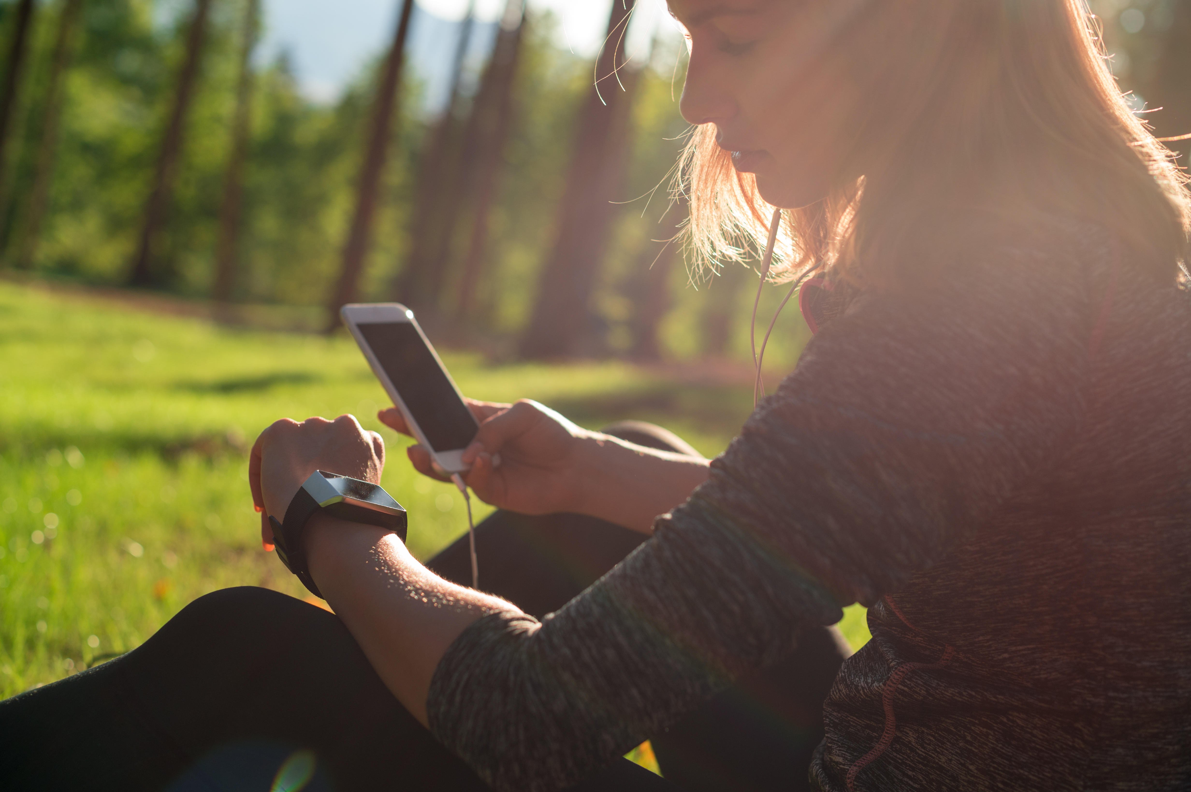 A photograph of a woman sat down on grass on a sunny day. In her right hand she's holding a mobile phone. She is looking at her smart watch on her left wrist. 