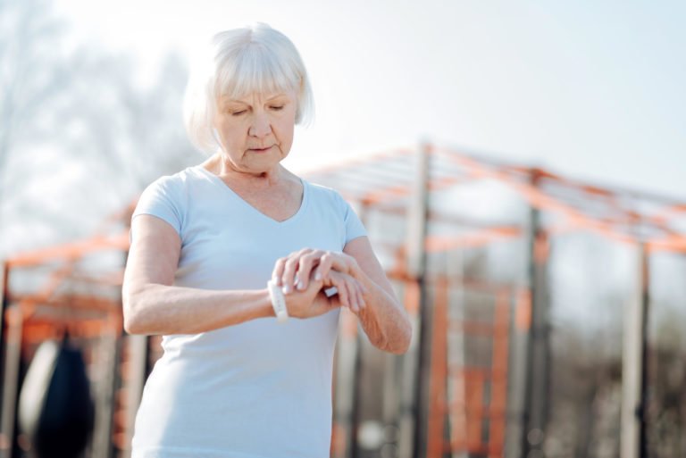 Concentrated older woman looking standing up outside looking at her fitness tracker. In the background out of focus is a large fitness apparatus.
