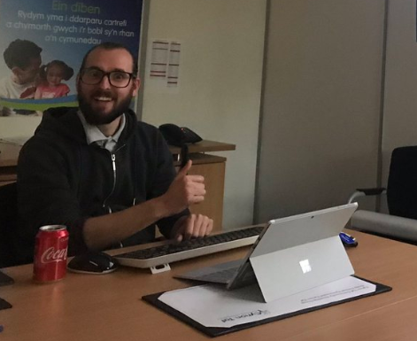 A photograph of Rhys Norris of Cynon Taff Housing at a digital inclusion training session. He is sat at a desk in front of a laptop. He smiles at the camera with a thumbs up.