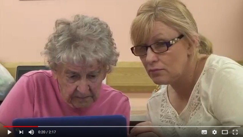 A screenshot from a YouTube video. A senior lady sits down looking at a tablet, while a female digital volunteer sits next to her, also looking at the tablet.