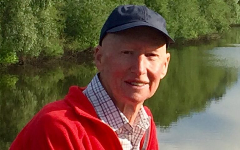 A portrait photograph of Peter Loughran, a Digital Volunteer. He is wearing a baseball cap. In the background is a river or a lake.