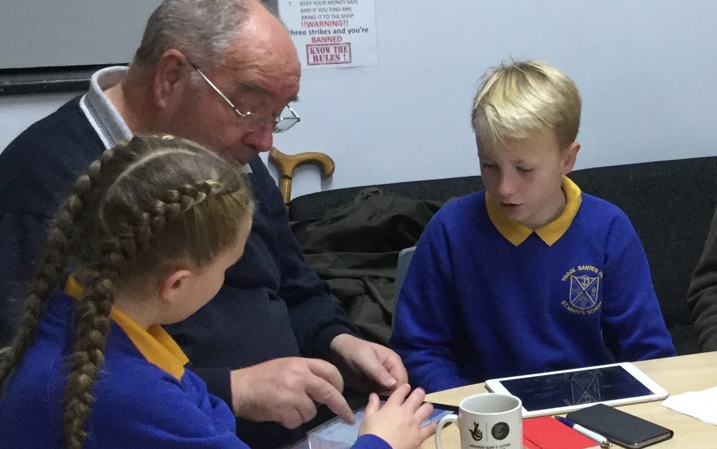 A photograph of a senior man sat at a desk being shown how to use a tablet by two school pupil digital champions. On the desk there is another tablet device. and two mobile phones.