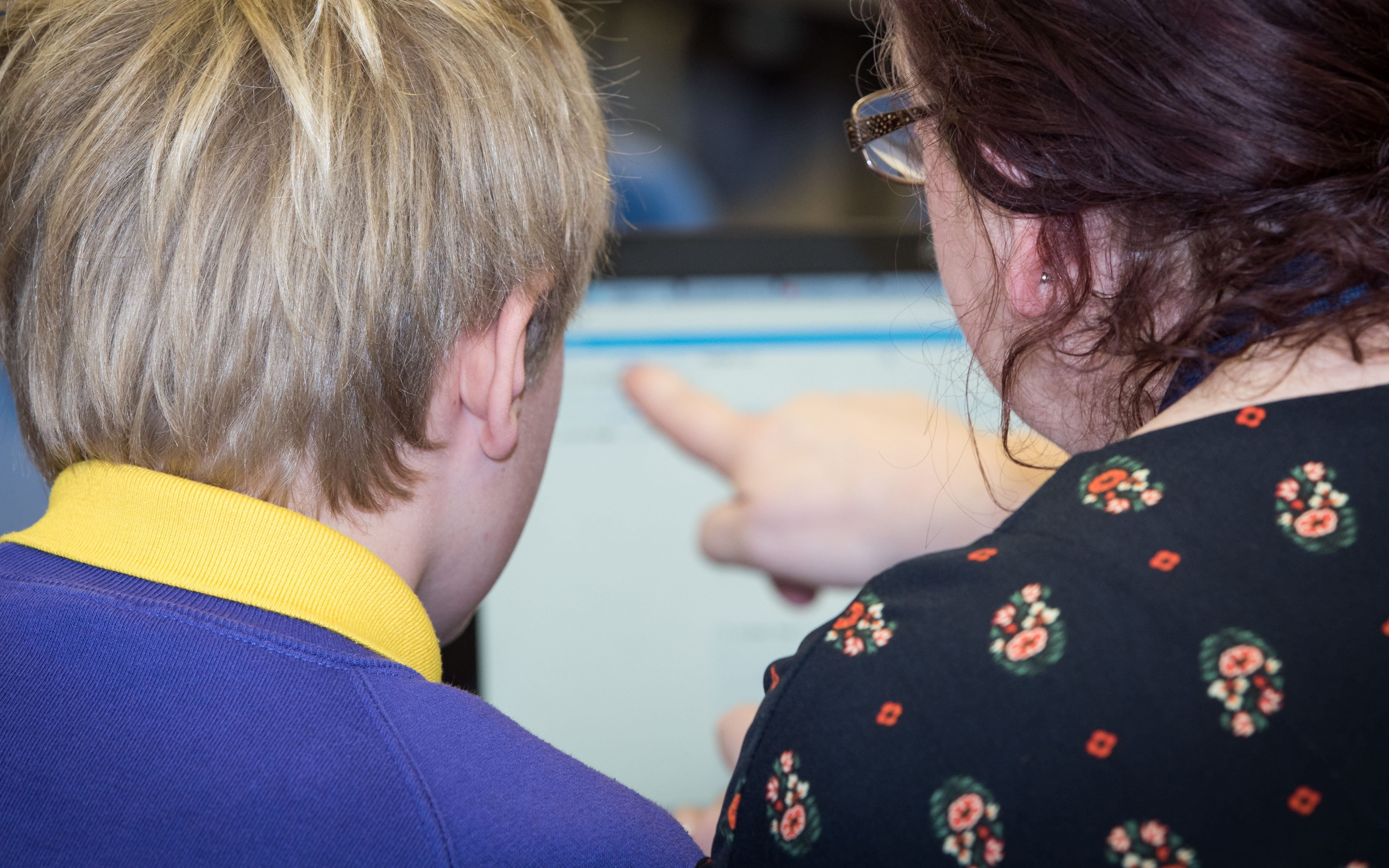 An over the shoulder photo of a teacher pointing at a computer screen. Sat next to her is a male pupil in school uniform.