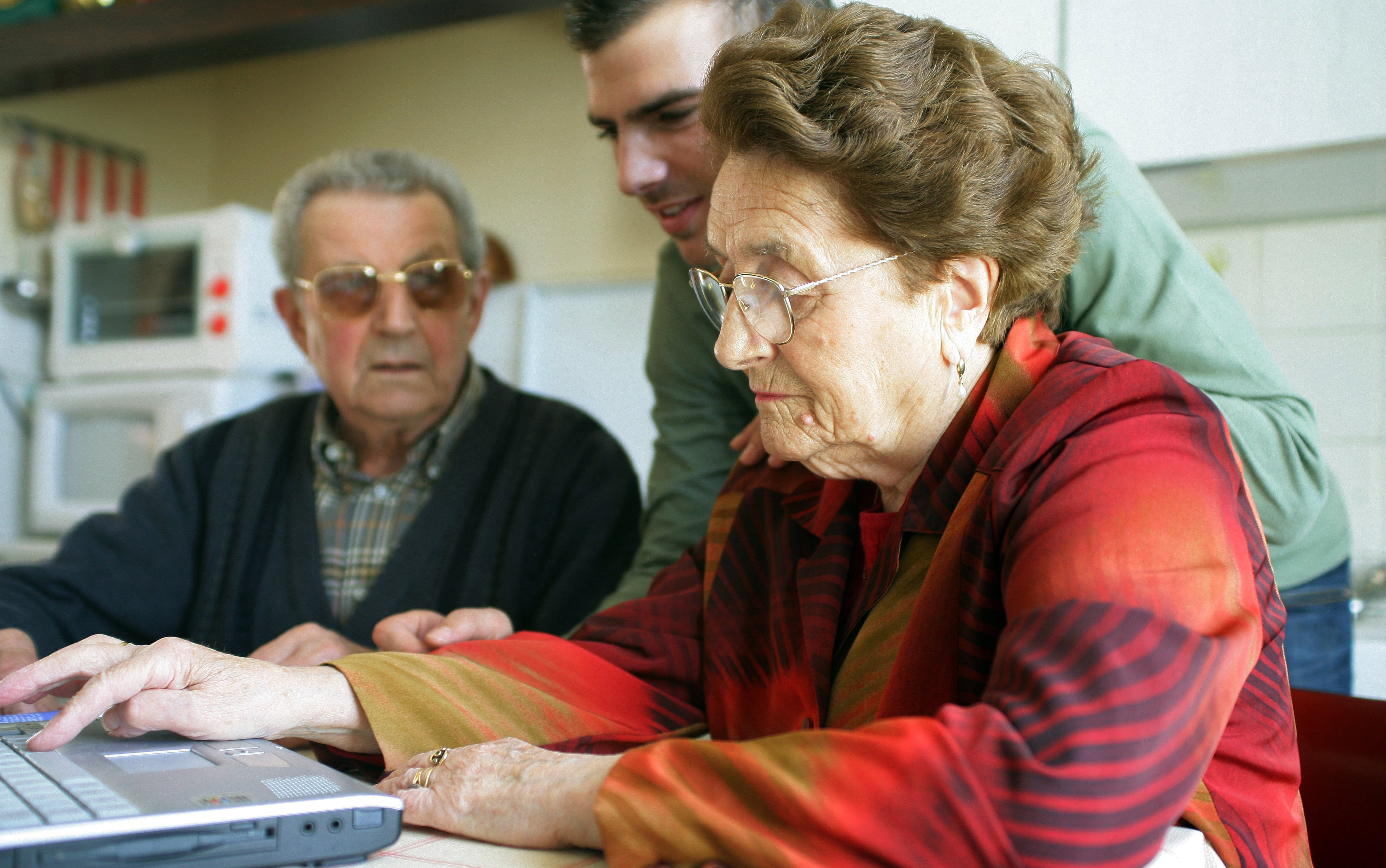 A photograph of a senior woman and a senior man sat down. They are being helped to use the laptop in front of them by a younger man standing in between them who is looking at the laptop screen.
