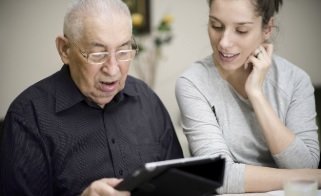 A photograph of a senior man sat down looking down holding a tablet device. There is a woman sat next to him also looking at the tablet, with her head resting on her raised left hand.