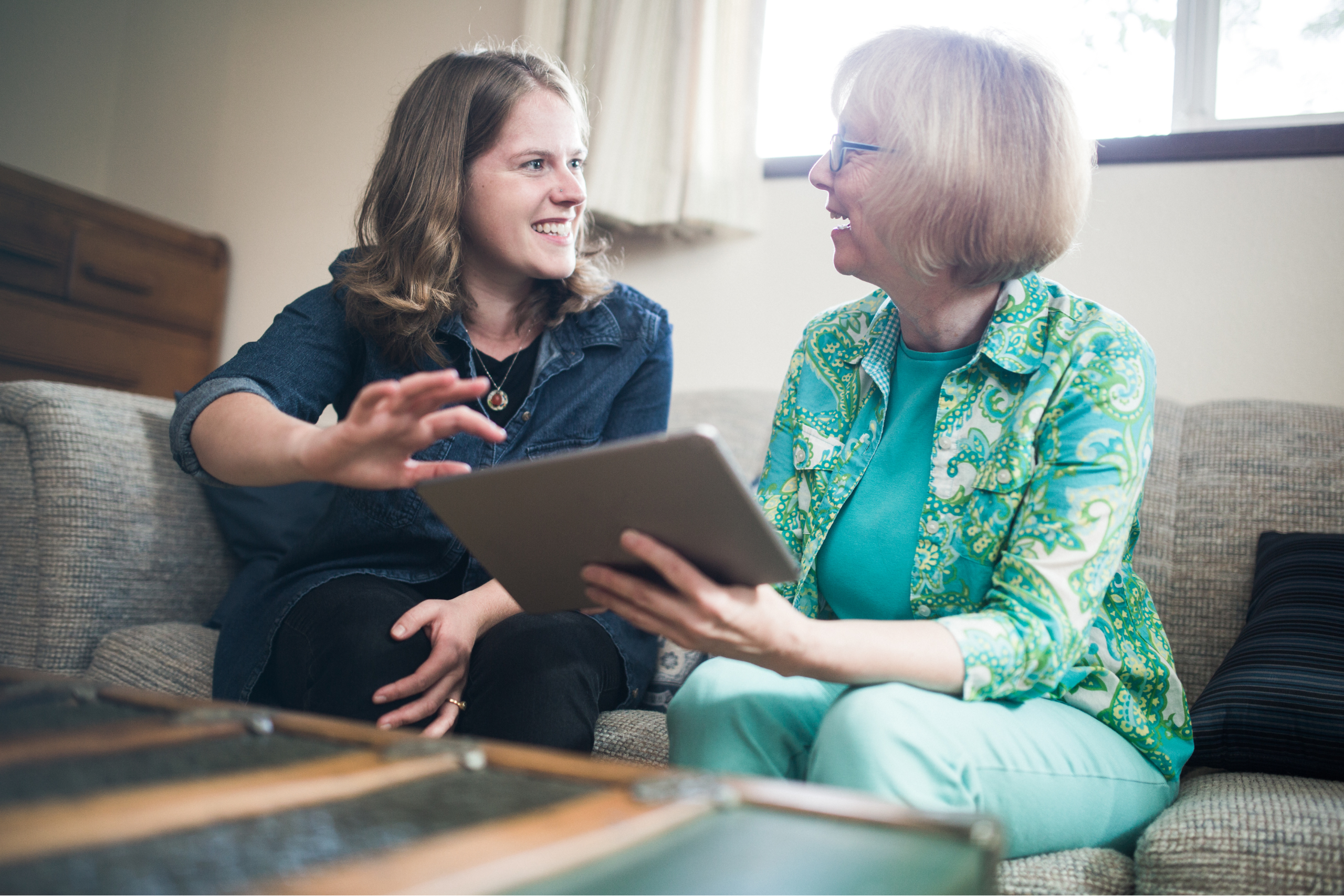 A photograph of two women sat on a sofa looking at each other and smiling. The older woman on the right of the photograph is holding a tablet device. The younger woman on the left points towards the tablet.