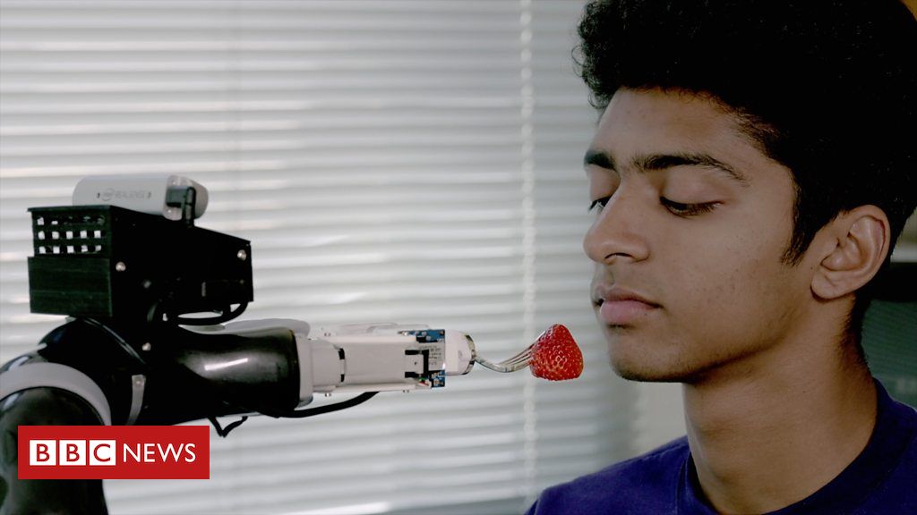 A photograph of a robotic arm holding a piece of food to an awaiting man who had his mouth closed and looking down at the food. In the bottom left corner of the image is the BBC logo.