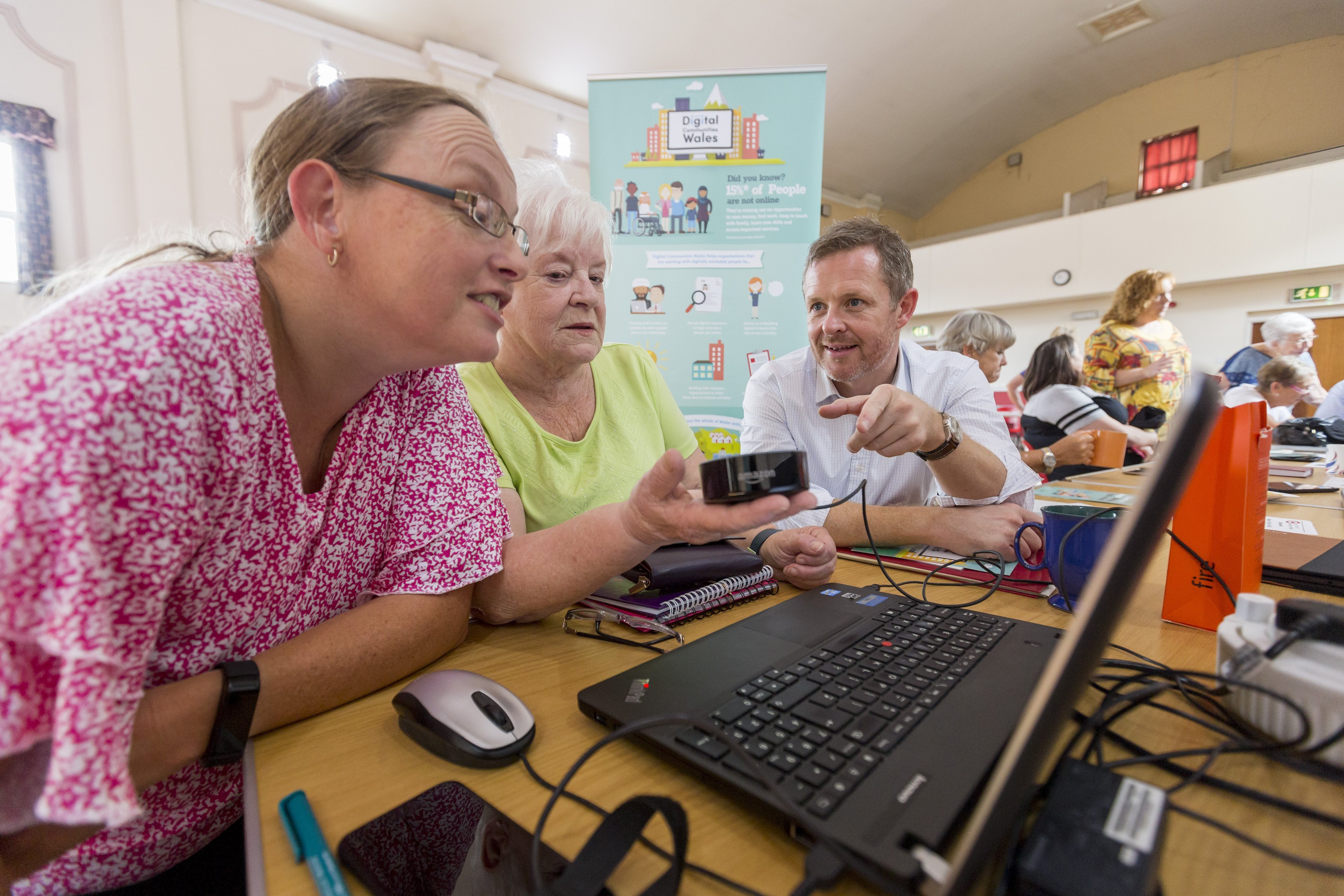 Senior woman holding an Amazon Echo sat in between a woman looking at a laptop and a man pointing at the Amazon Echo