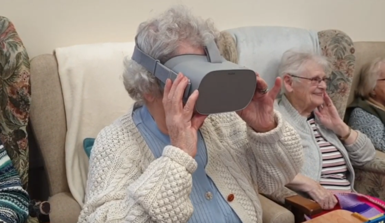 Photograph of a senior lady sat down using a VR headset at Age Well Bontnewydd. In the background there is another senior lady sat down smiling.