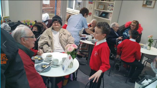 A photograph of Ysgol Emmanuel e-cafe. The image shows a room full of people mostly sat down around circular tables and chatting. In the foreground there is two older men sat around a table talking to a pupil who is standing.