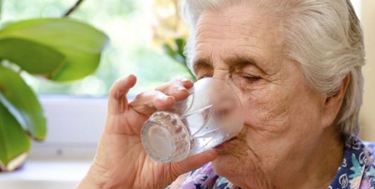 A closeup photograph of a senior lady drinking a glass of water. In the background is a window, leaves of a plant and a flower.