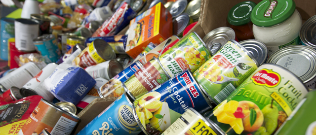 A close up photograph of tins, jars and boxes at a foodbank. Foods include, tinned fruit and vegetables, cooking sauces, sugar and rice.
