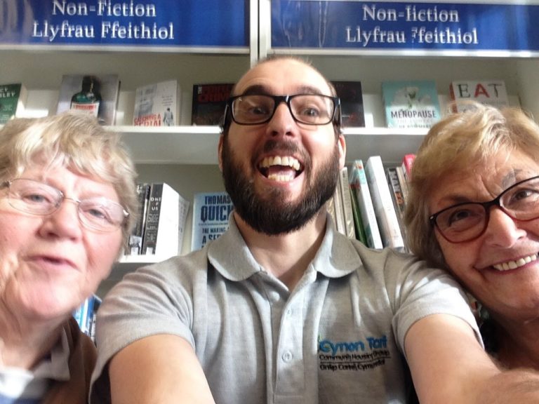 A selfie photograph of three people. In the middle is Rhys Norris of Cynon Taff Housing smiling at the camera. Either side of him are two senior women whole are also smiling. In the background are shelves of library books, the sign above the books reads 'Non-Fiction, Llyfrau Ffeithiol'