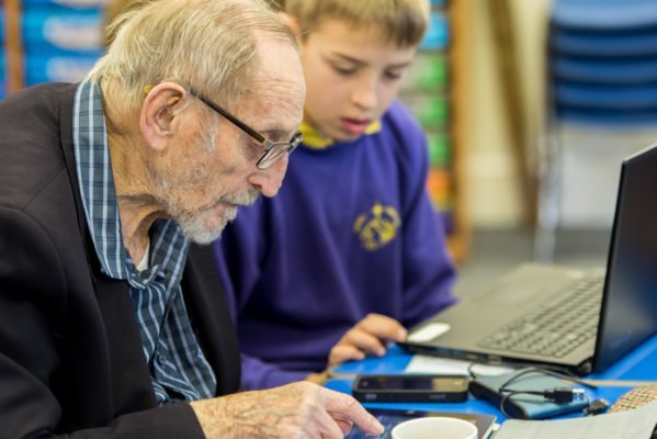 Senior man looking at a mobile phone on a table next to a young boy in school uniform sat in front of a laptop