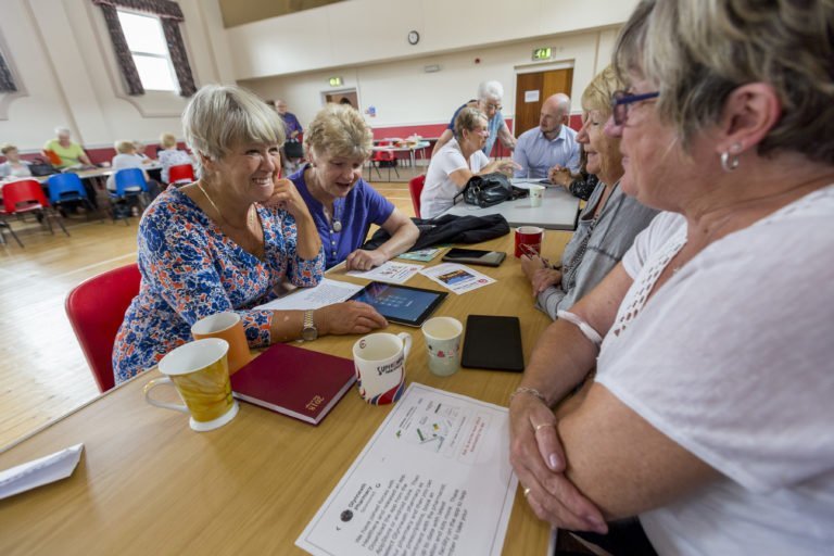 Senior ladies sat either side of a long desk chatting. On the desk is a tablet device, coffee cups, a diary planner and paper documents.