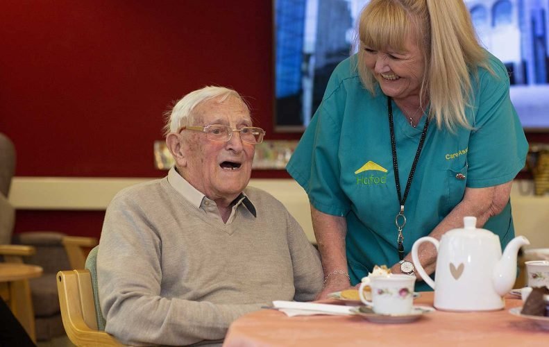 A photograph of a Ty Gwyn resident, who is an older man sat down behind a table, and staff member, who is standing next to the older man and smiling. On the table there is a teapot and a teacup.