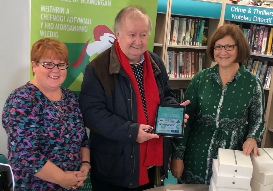 A photo from the Vale Tablet Loan Scheme launch. In the image is two older women stood either side of an older man in a library, all are smiling. The man is holding a tablet device.