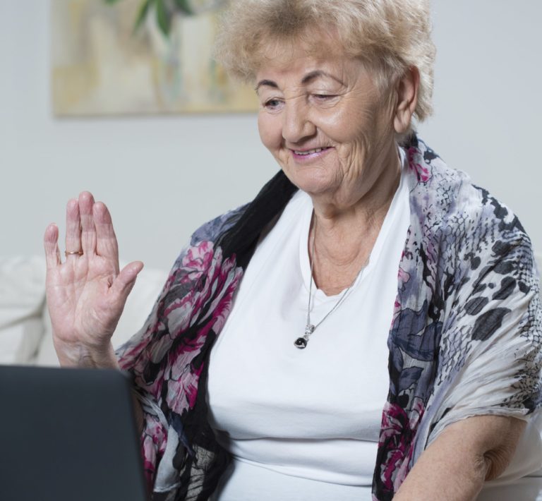 Photo of elderly woman sat down with her hand raised in a 'hello' gesture, looking at a laptop, suggesting that she is on a video call.