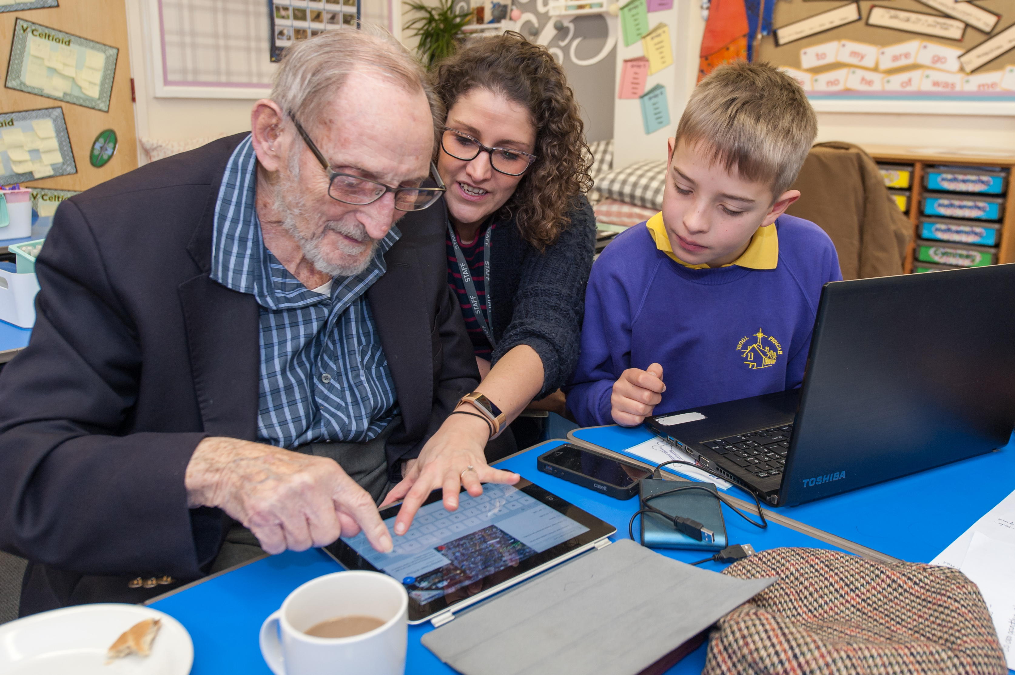 A photograph of a female teacher and a male primary aged pupil teaching a senior man to use an iPad over a cup of tea. In front of the pupil there is an open laptop.