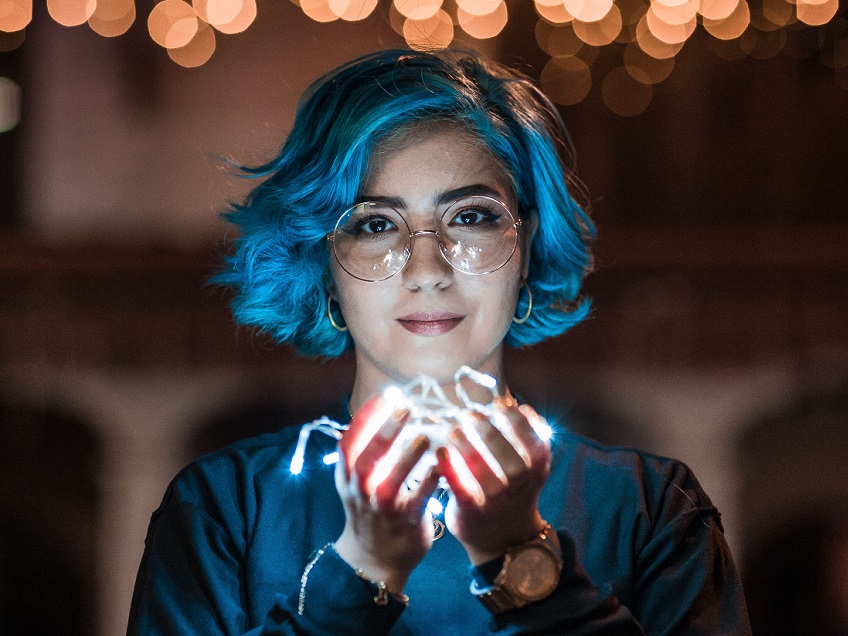 A photograph of a young woman with blue hair and glasses holding a string of LED lights in a ball in front of her. The glow from the lights illuminates her face.