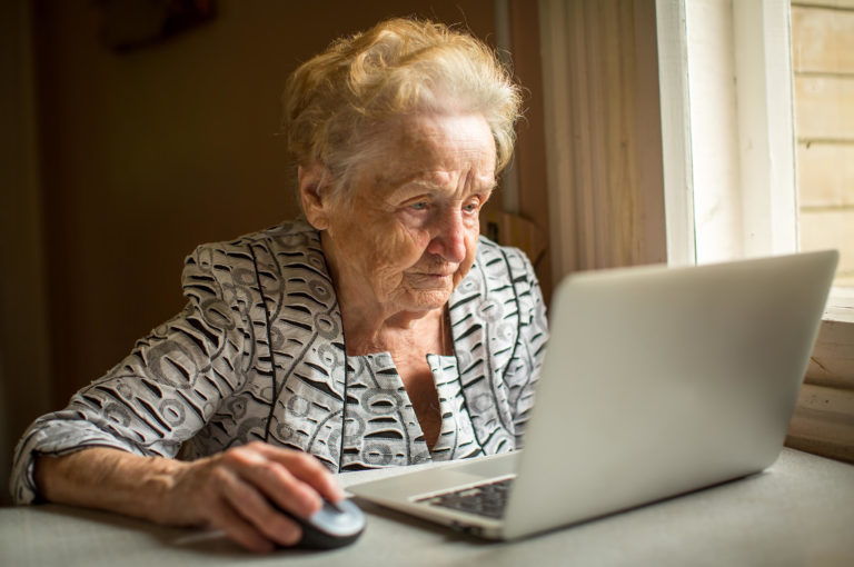 A photograph of an elderly woman sat at a desk using a laptop with a computer mouse.