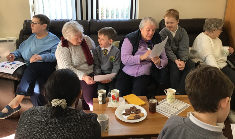 Digital Heroes from Ysgol Borras meet residents from Cae Glo. In the foreground of the photo is the back of the heads of two pupils in school uniform. In the background of the photo are four senior ladies and two children all sat on a sofa and interacting with one another. On the table in the middle of the photograph there are teacups and a plate of biscuits.