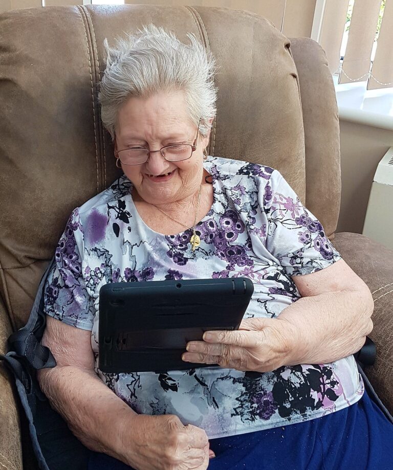 A photograph of an elderly woman sat in an armchair smiling while looking at a tablet that she is holding.