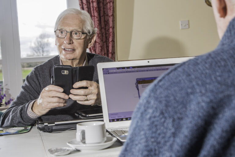 An over the shoulder photograph of a man almost out of shot sat down and looking at a laptop screen. In the background of the photo the main subject is a senior man looking into the distance holding a mobile phone with both hands.
