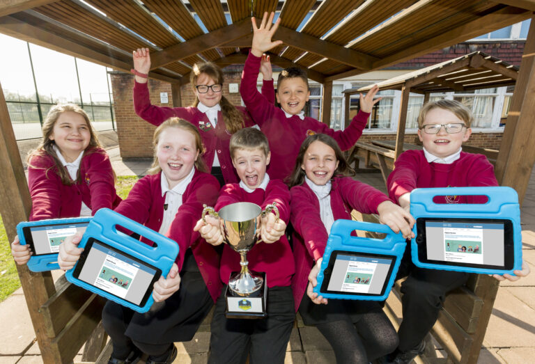 A photograph of Digital Heroes at St Julians School. There are two pupils at the back with their hands in the air. There are five pupils in the front. The middle pupil is holding a trophy and the four either side are holding tablets.
