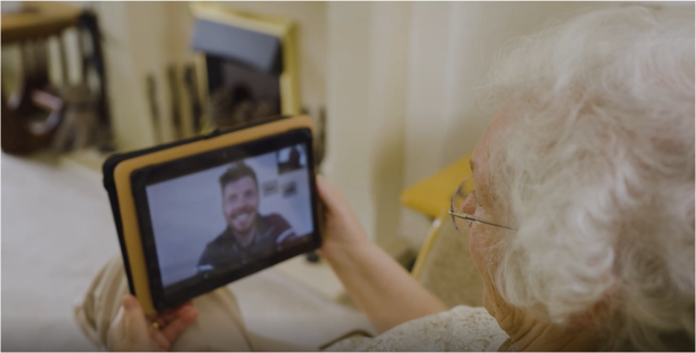 An over the shoulder photograph of a senior lady making video call on tablet device. There is a smiling younger man on the screen, who she is talking to.