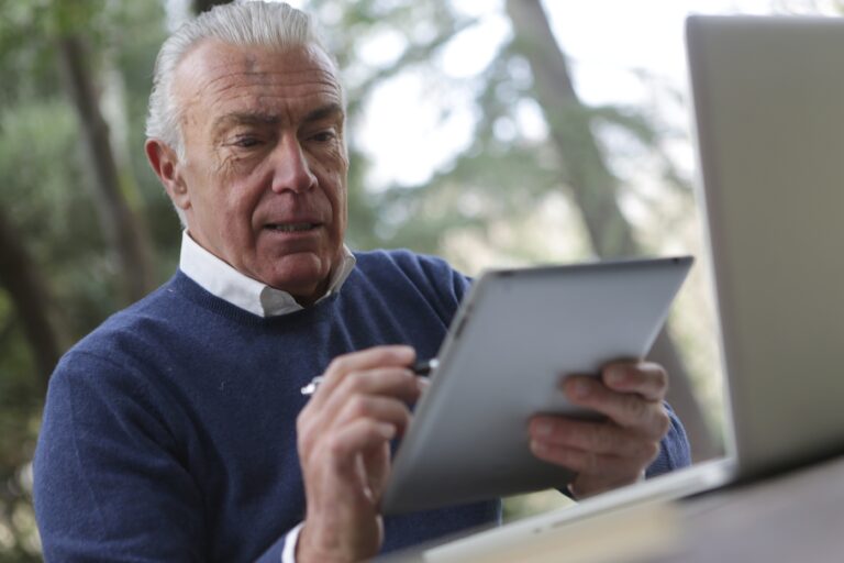 A photograph of a senior man using a stylus to interact with a tablet device. He is sat at a desk with a laptop open in front of him.