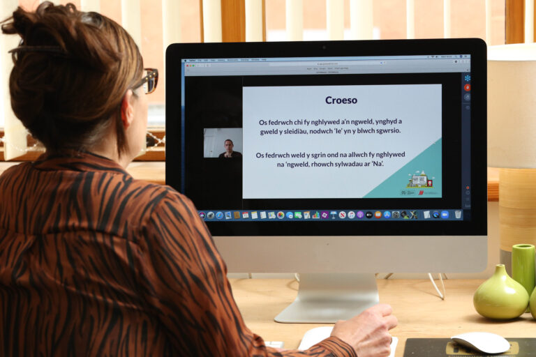 An over the shoulder photograph of a woman sat down using a desktop computer. She is watching a DCW webinar on the screen.