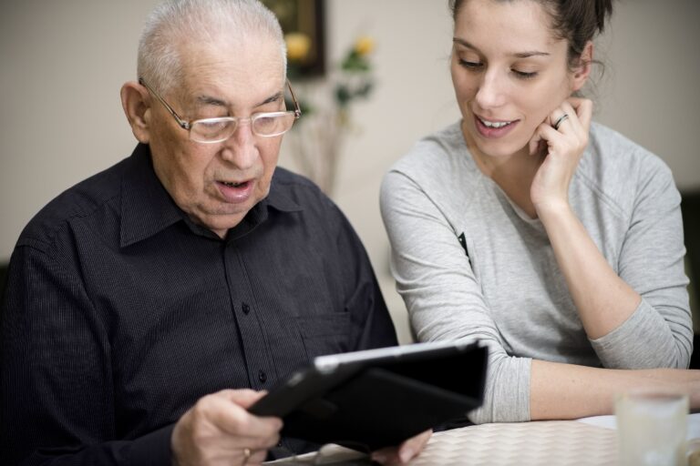 A photograph of a senior man and a younger female digital champion. The senior man is sat looking down at the tablet device he's holding. The younger female digital champion is looking at the tablet with her head resting on her left hand.