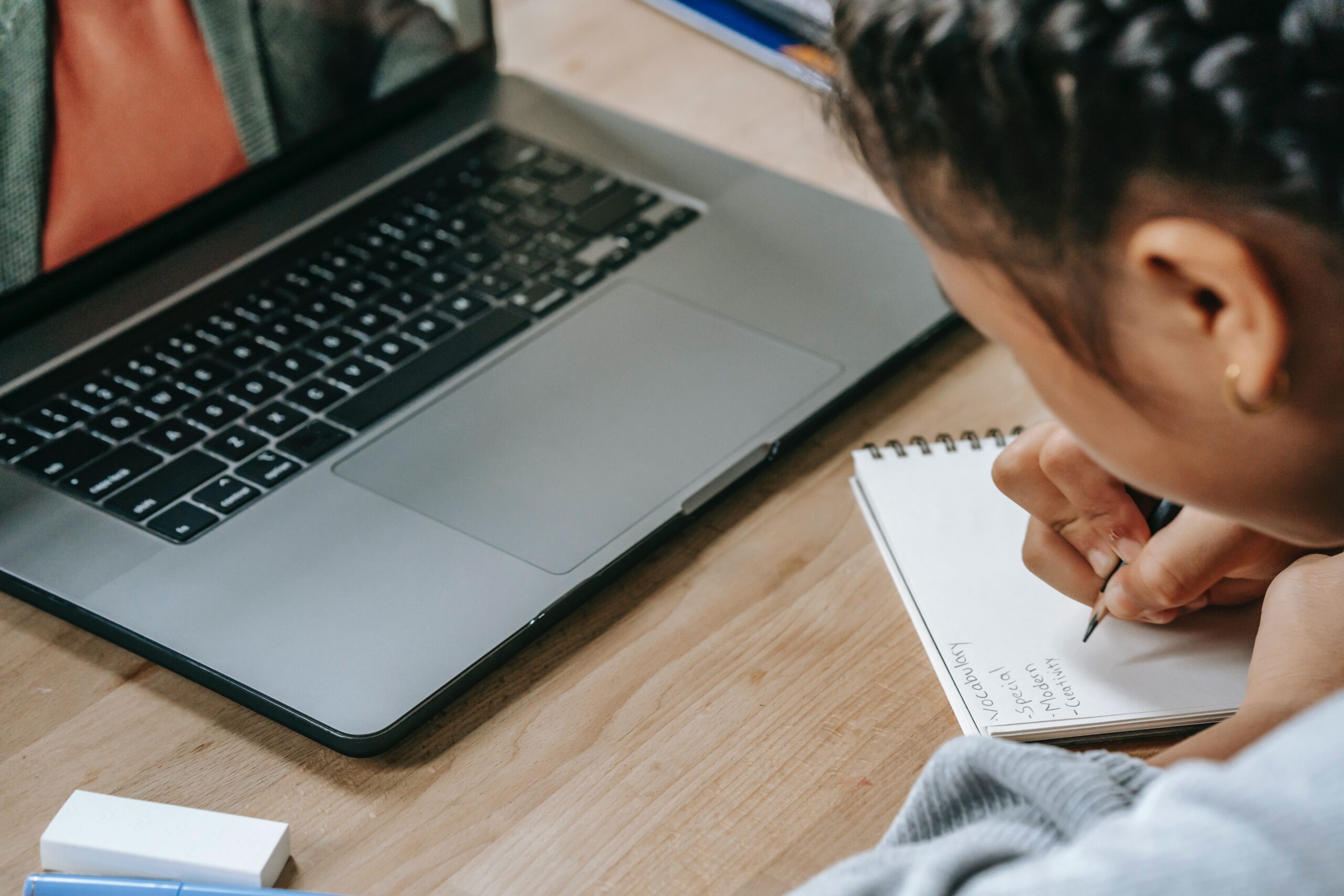 An over the shoulder photograph of a young girl using laptop for homework while writing on a notepad.