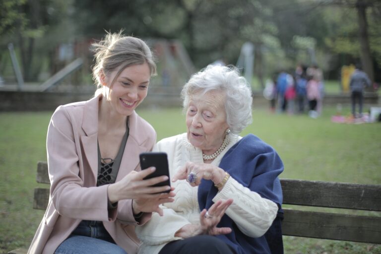A photograph of a young woman showing a senior woman how to use a mobile phone. They are both sat on a bench at a park.