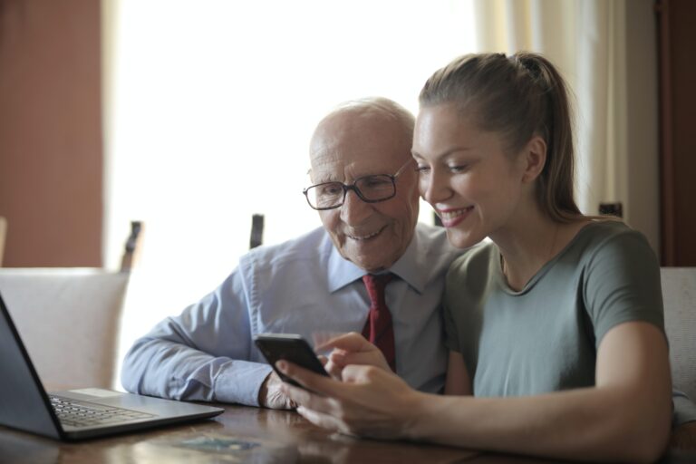 A photograph of a young woman helps an elderly man use a mobile phone. They are both sat down at a desk with a laptop on it.